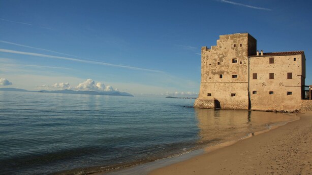A coastal building with a tower, situated by a calm sea with clear skies.