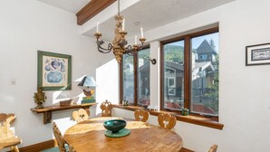 A dining room with a wooden table, chairs, a chandelier, and large windows with a view of houses. - Plaza Lodge | 3 - 2 Bedroom Silver (Vail)