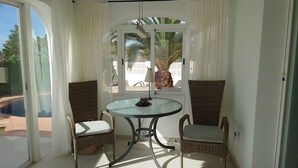 A sunlit room with a glass table, two wicker chairs, and a view of a swimming pool and palm trees outside. - Casa Zinat para 4 Personas (Calpe)