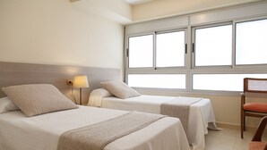 Une chambre d’hôtel avec deux lits, une fenêtre avec des stores, une chaise et une table de chevet avec une lampe.