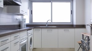 Une cuisine moderne avec des meubles blancs, un four encastré, un lave-linge et une grande fenêtre qui laisse entrer la lumière naturelle.