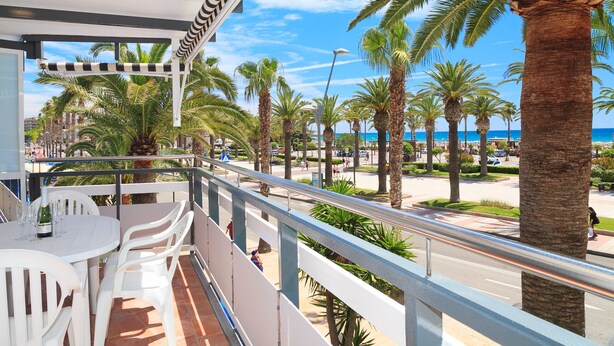 Un balcon avec une table et des chaises blanches, donnant sur une rue bordée de palmiers et sur la plage.