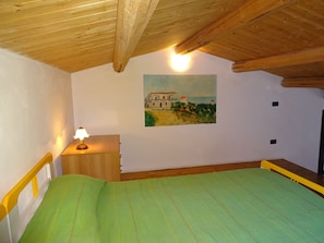 Une chambre avec un plafond en bois, un lit recouvert d’une couverture verte, une table de chevet avec une lampe et un tableau représentant une maison accroché au mur.