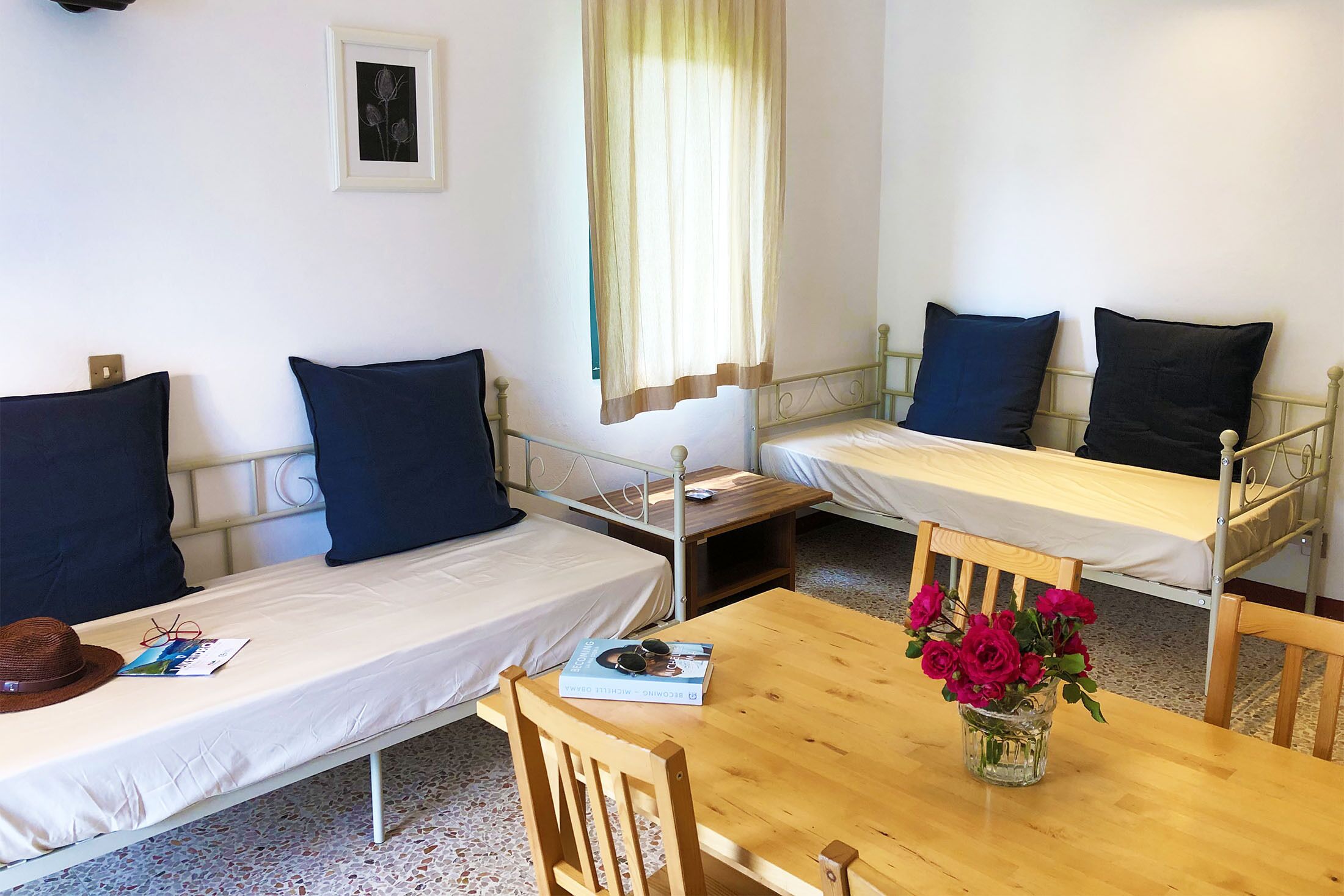 A room with two single beds, a wooden table, a chair, a vase with flowers, a framed picture on the wall, and a window with curtains.