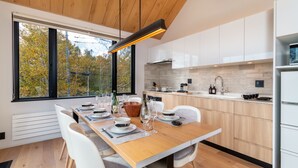A modern kitchen with white cabinets, a wooden dining table set for four, and a large window overlooking a forest. - Koa Niseko (Niseko)