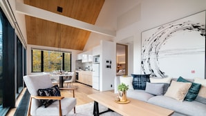 A modern living room with a sofa, a coffee table, and a chair. There is a kitchen in the background with cabinets and a dining area. - Koa Niseko (Niseko)