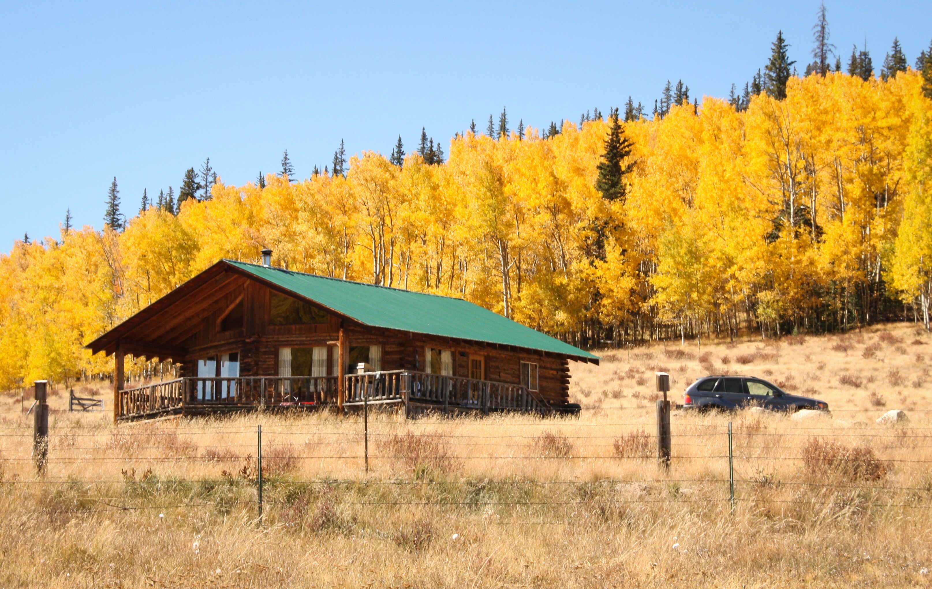 A rustic cabin with a green roof, surrounded by a wooden fence, set against a backdrop of yellow autumn trees.