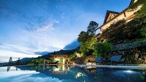 A serene infinity pool reflecting a traditional building and lush greenery, surrounded by stone walls and lounge chairs. - Beautiful architect villa overlooking the lagoon and the island of Tahiti (Te'avaro)
