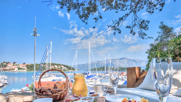 A table set with food and drinks, overlooking a marina with boats and houses in the background. - Eco Aparthotel The Dreamers' Club - Garden for 4 (Lumbarda)