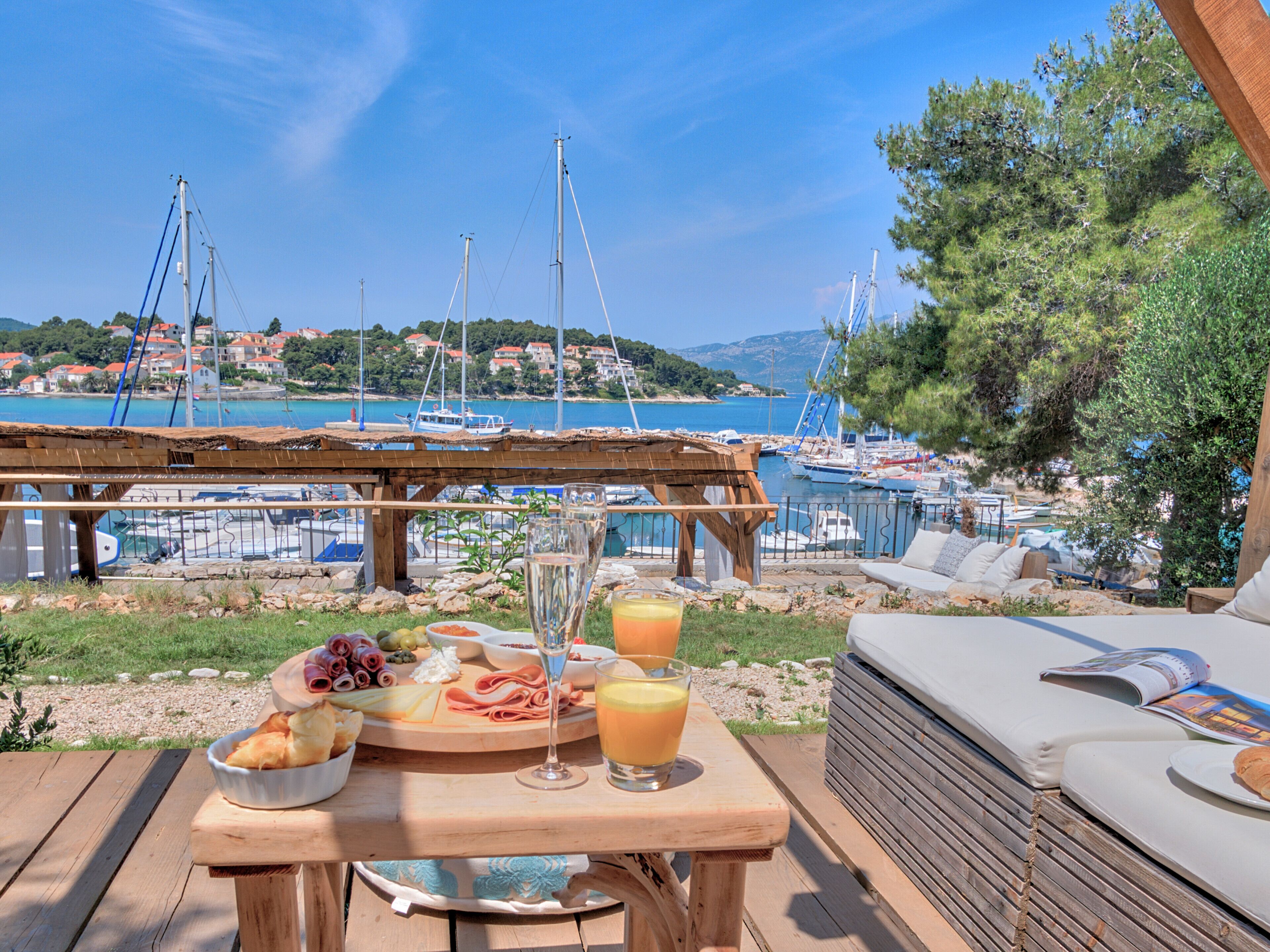 A wooden table with a meal and drinks, overlooking a marina with boats and a coastal town.