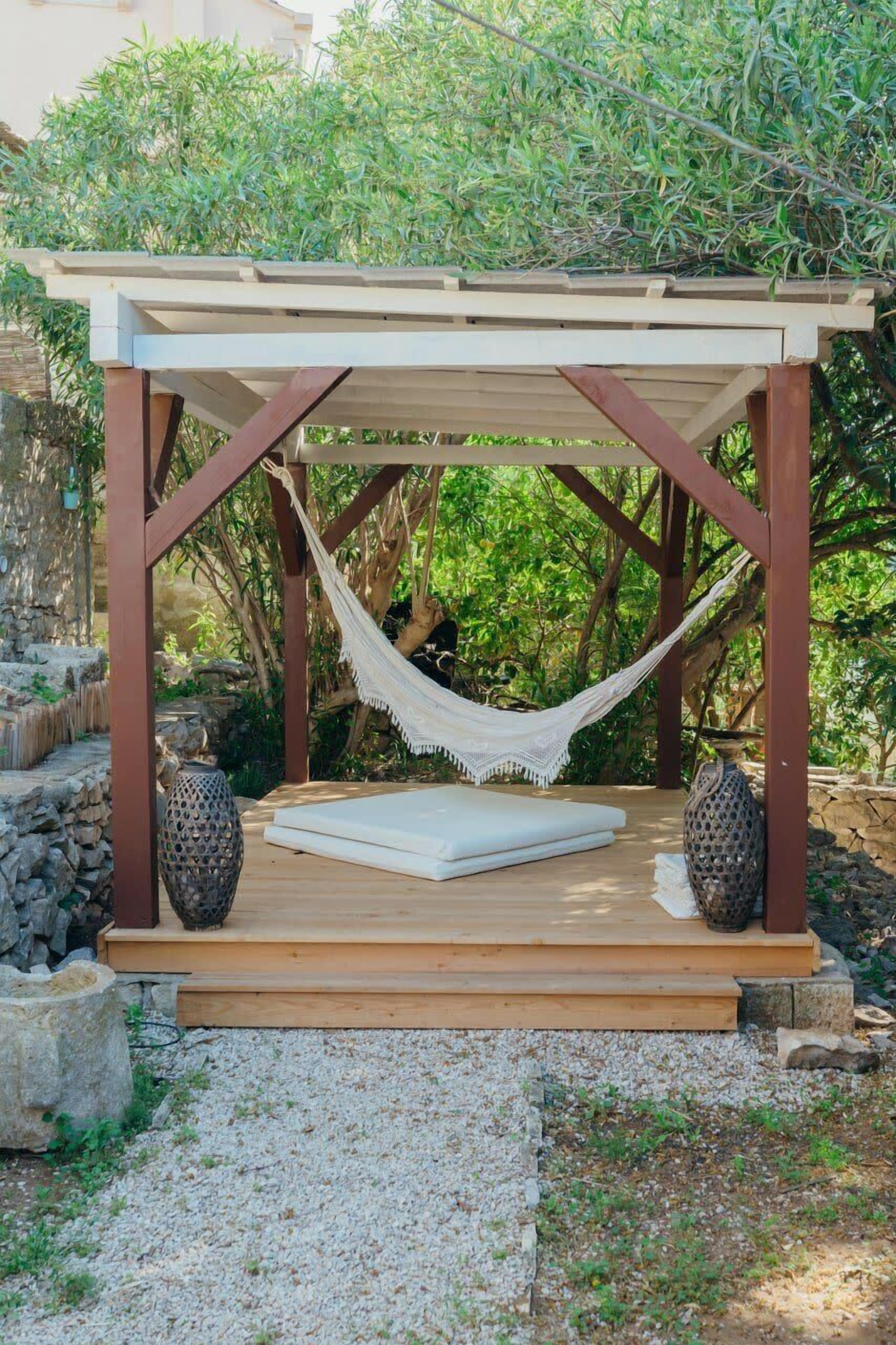 Un espace extérieur aménagé en bois, avec un hamac, un banc rembourré et des vases décoratifs.