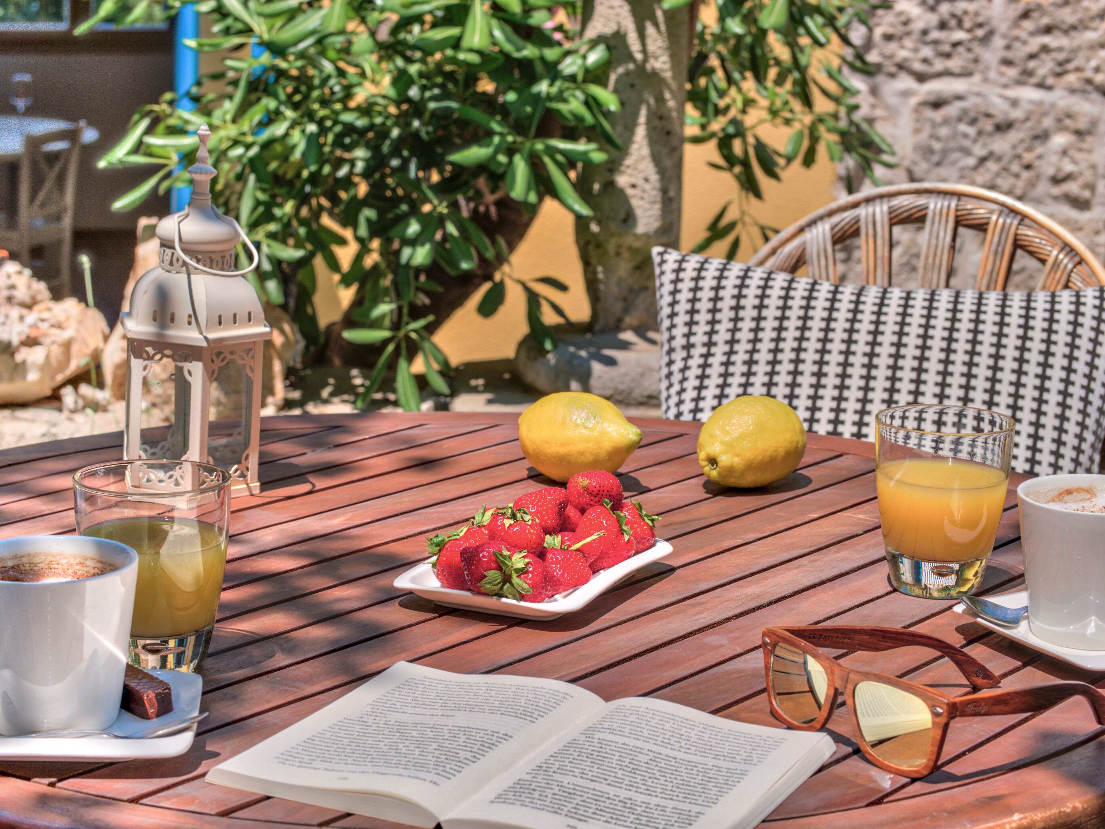 Une table en bois sur laquelle se trouvent un livre, une assiette de fraises, des citrons, un verre de jus d’orange, une tasse de café et une paire de lunettes de soleil.