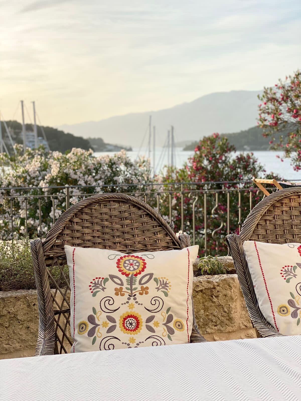 Wicker chairs with floral cushions and a table, set against a scenic backdrop of a body of water and mountains.