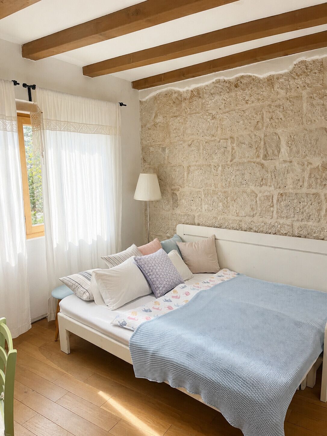A bedroom with a stone wall, a bed with a blue blanket, and a window with white curtains.
