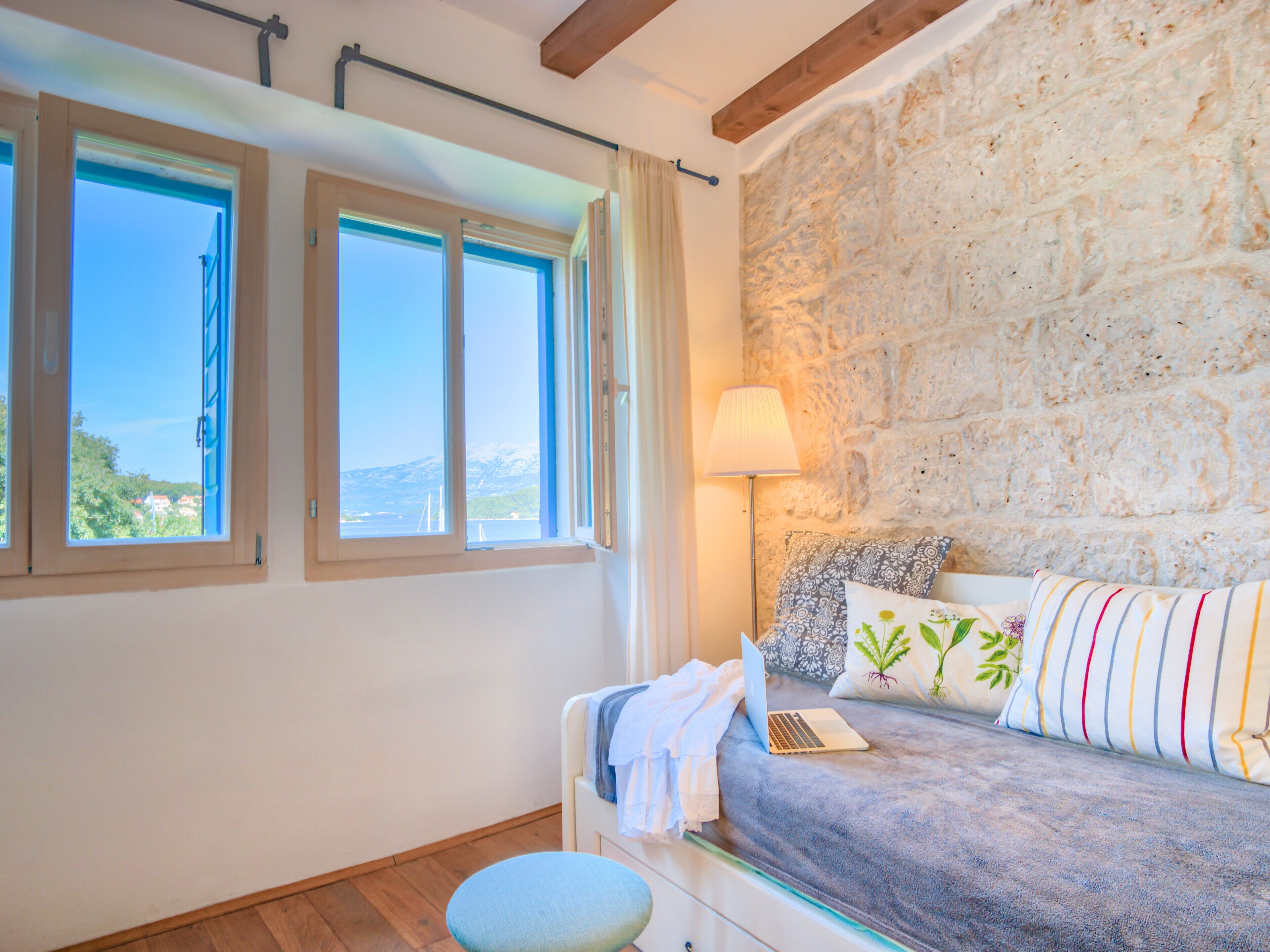 Quarto com parede de pedra, uma cama com um laptop, uma luminária e vista para o mar através de grandes janelas.