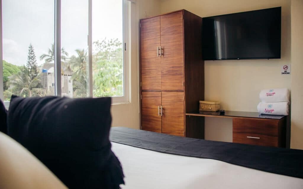 A hotel room with a bed, a wooden wardrobe, a flat-screen TV, and a window with a view of greenery.
