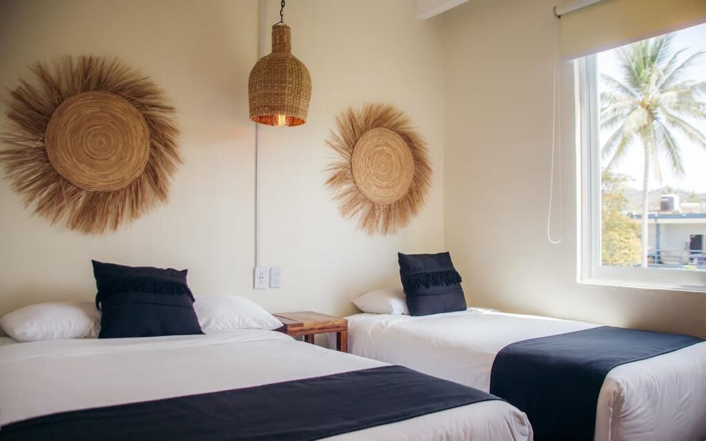 Habitación de hotel con dos camas, una lámpara colgante y ventana con vistas a palmeras.