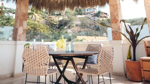 An outdoor patio with a thatched roof, stone pillars, wicker furniture, and a view of the surrounding landscape. - Nuiya Hoteles (Sayulita)