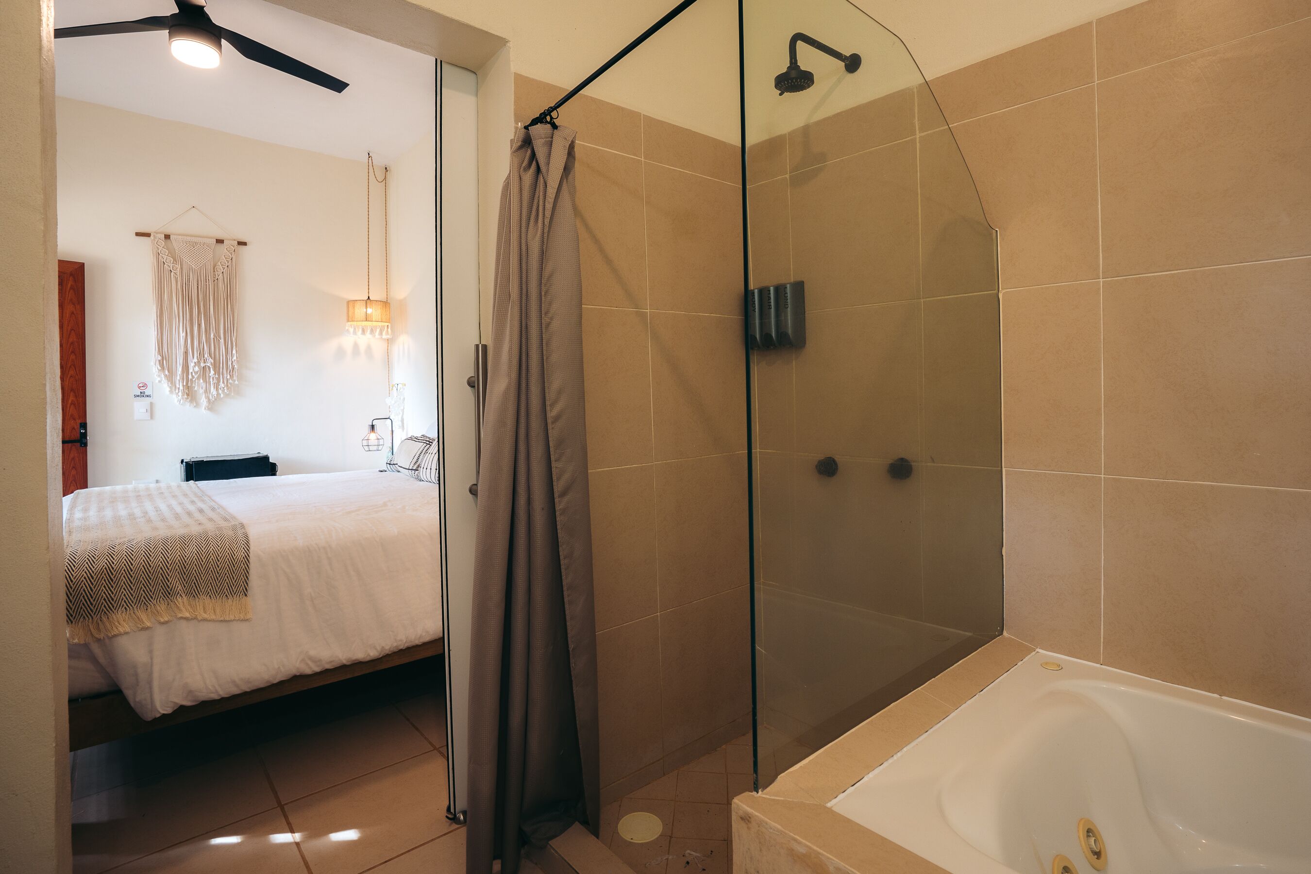 A modern bathroom with a glass-enclosed shower, a bathtub, and a visible bed with a white bedspread and a beige throw blanket.