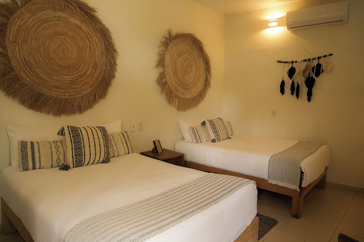 Deux lits avec du linge de lit blanc et des oreillers à motifs, un climatiseur fixé au mur et une décoration murale composée d’objets suspendus.
