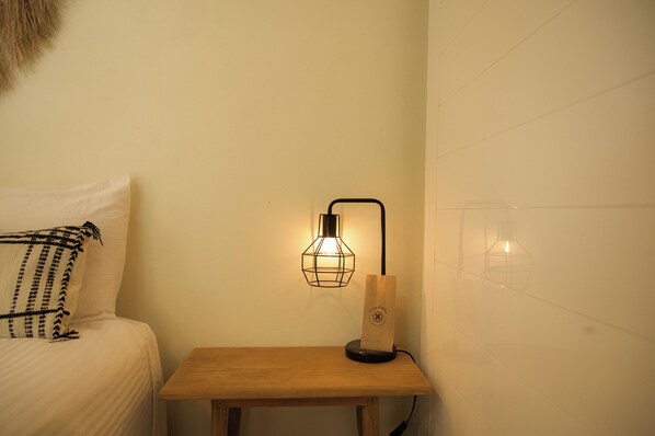A neatly made bed with a patterned pillow, a small wooden nightstand with a lamp, and a wall socket.
