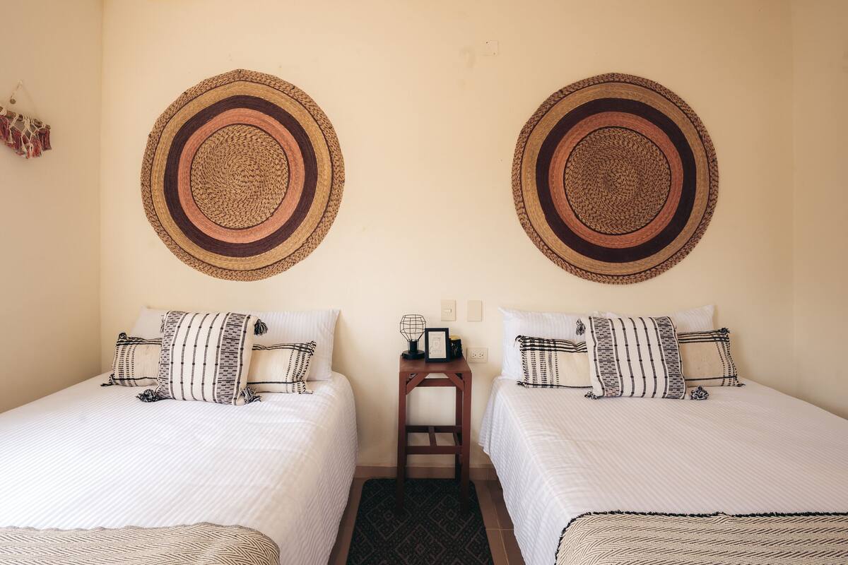 Une chambre avec deux lits, chacun agrémenté d’oreillers à rayures et d’une tête de lit en osier. Une petite table de chevet en bois, sur laquelle se trouvent une lampe et un tableau encadré, est placée entre les deux lits.