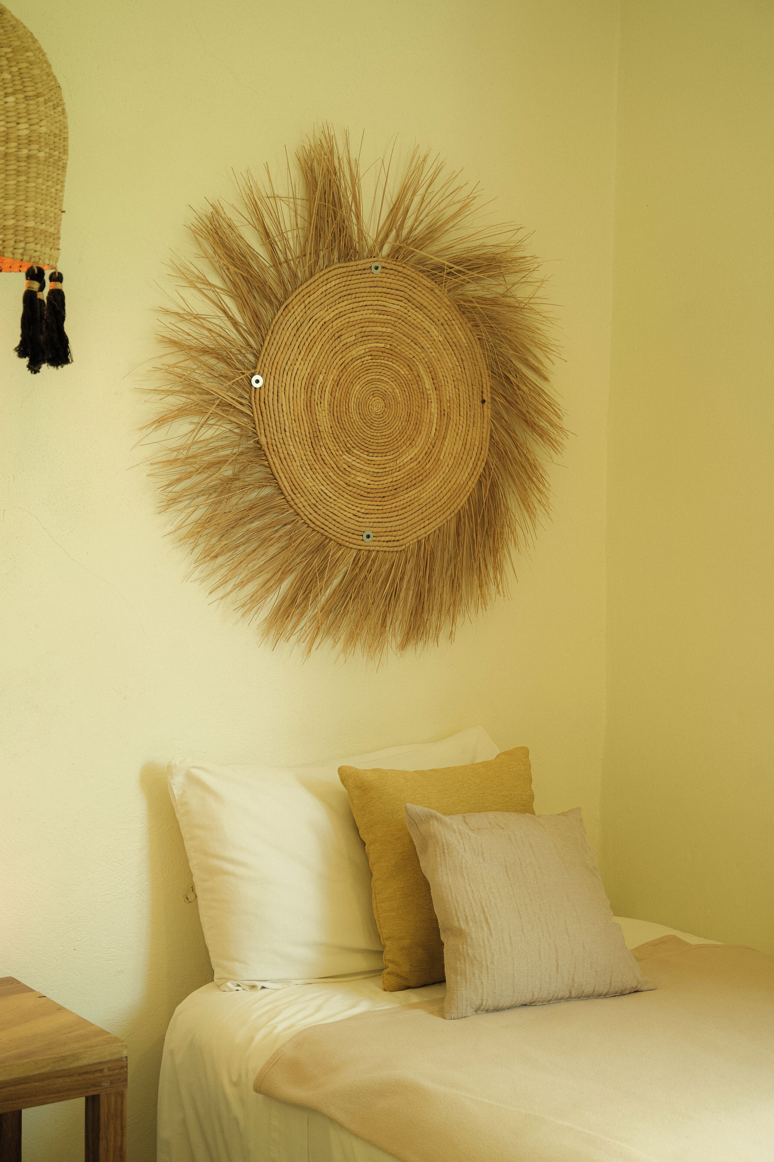 Una cama con ropa de cama blanca y beige, un adorno circular tejido en la pared y una pequeña mesa de madera a su lado.