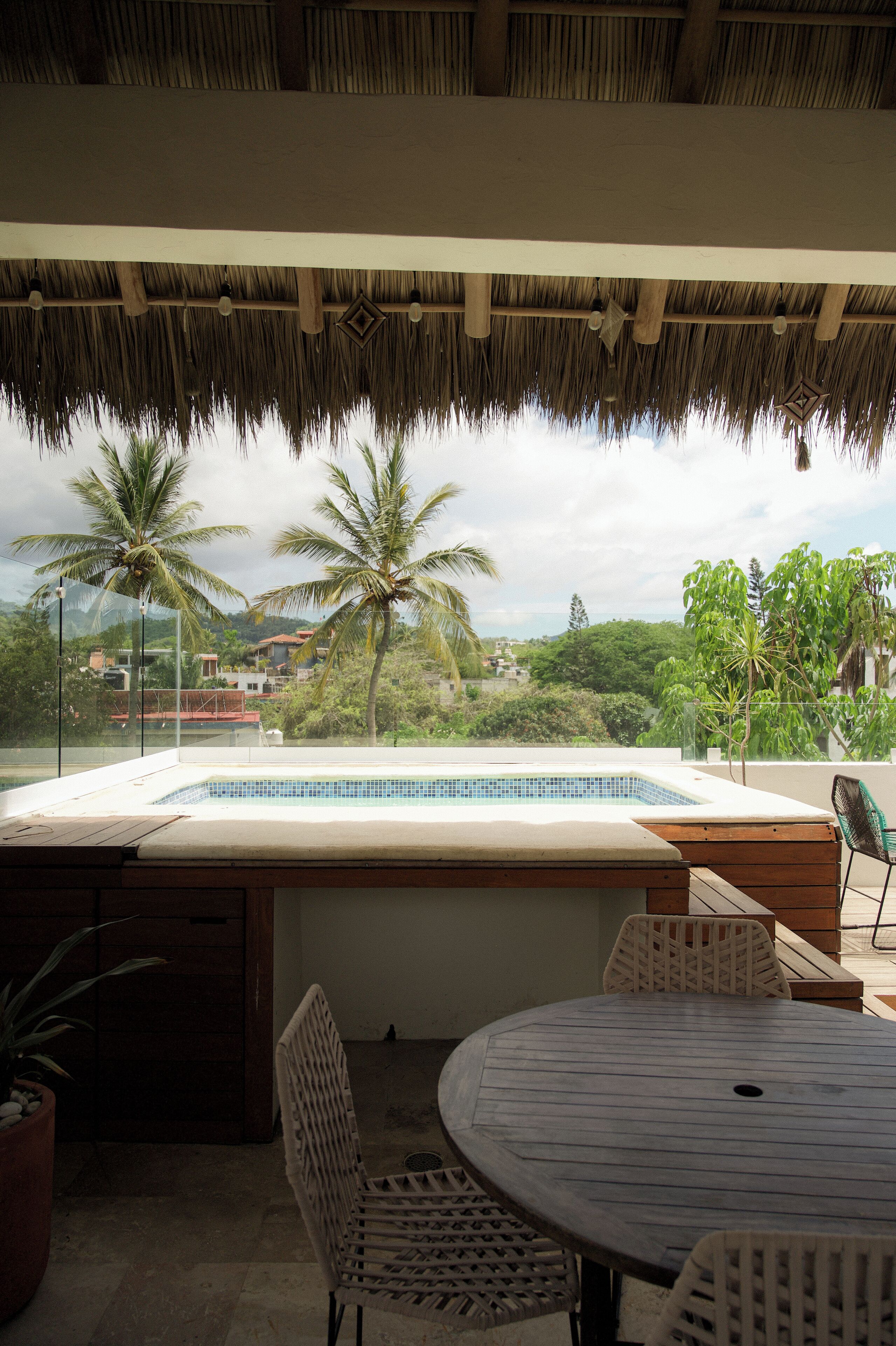 Zona de piscina con techo de paja, una mesa redonda y sillas, rodeada de plantas en macetas y con vistas panorámicas a palmeras y edificios.