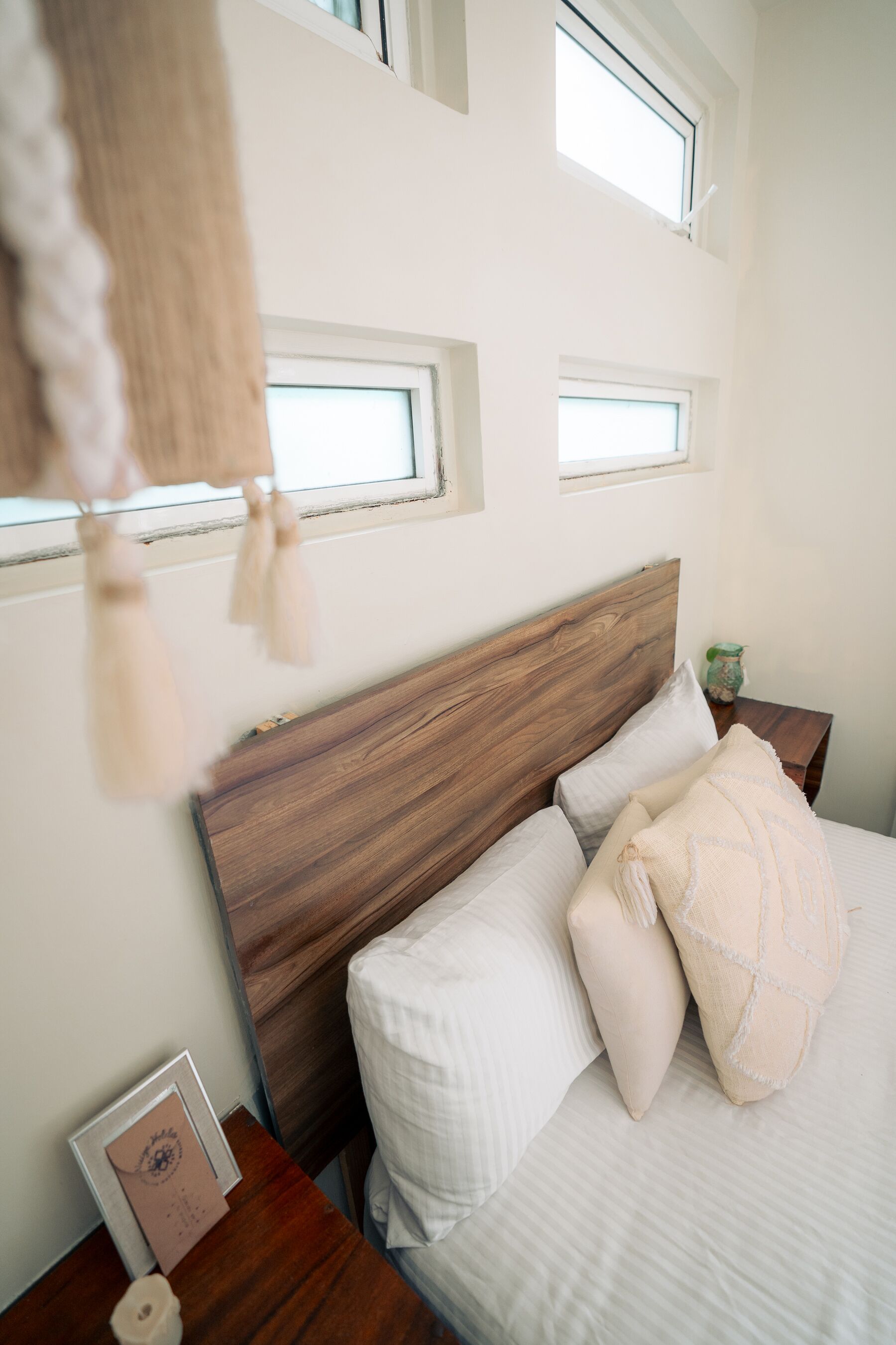 A bedroom with a wooden headboard, a bed with white bedding, and a nightstand with a framed picture and a small vase.