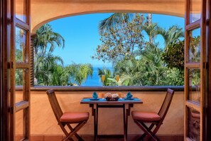 Enjoy a tranquil retreat with a view of the tropical landscape from the comfort of this room, featuring a table for two and a refreshing beverage.
