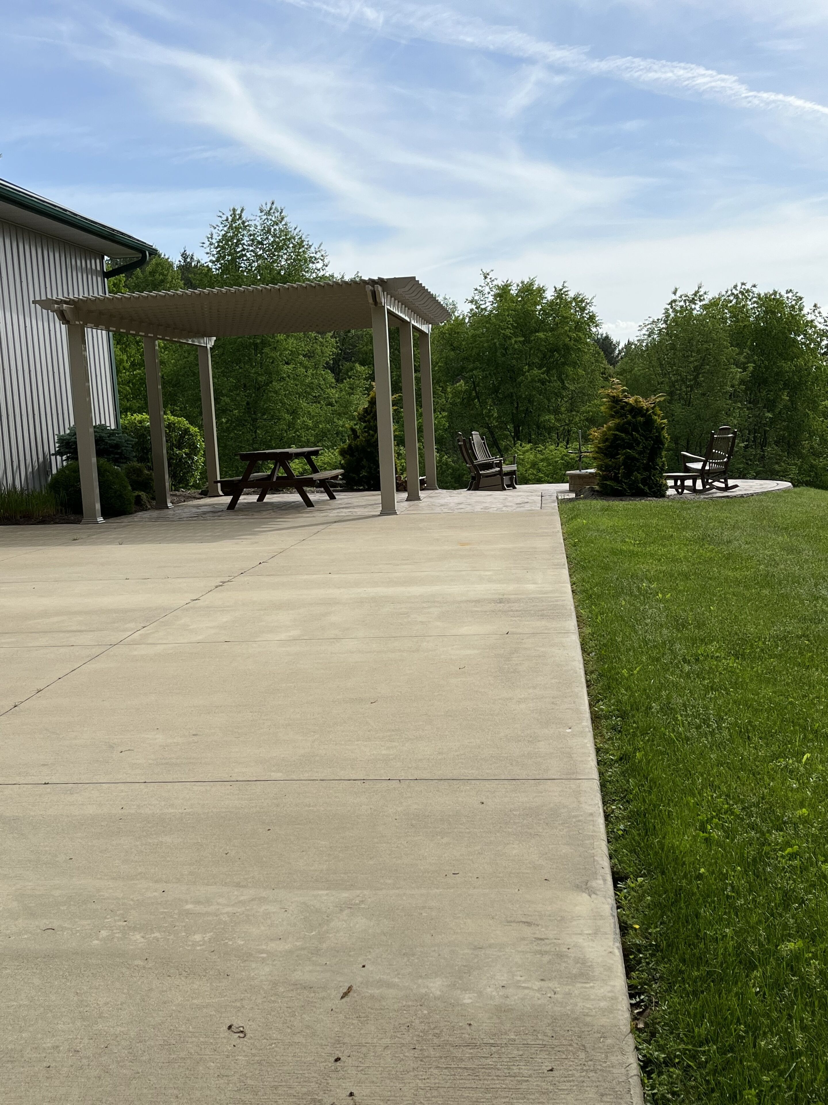 Une terrasse en béton avec des tables de pique-nique et une pergola, entourée d’une pelouse et d’arbres.