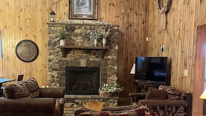 Un salon chaleureux avec une cheminée en pierre, des murs en bois, un ventilateur de plafond et un téléviseur.