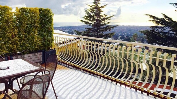 Un balcone con tavolo e sedie, un pavimento ricoperto di neve e vista sulla città e sugli alberi.