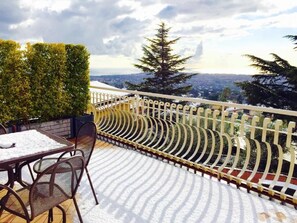A balcony with a table and chairs, a snow-covered floor, and a view of a cityscape and trees.