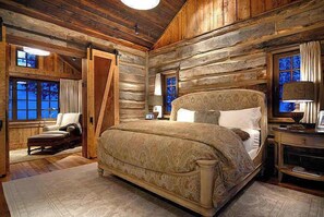 A log cabin bedroom with a large bed, a chair, a desk, and a window with curtains. - Tipple House (Mt. Crested Butte)