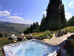 A hot tub with outdoor seating, surrounded by a stone patio and a scenic view of hills and trees. - Tipple House (Mt. Crested Butte)
