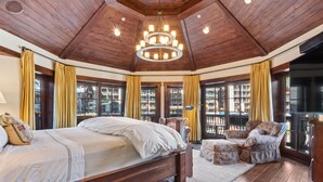 Um quarto espaçoso com uma cama grande, um lustre, teto de madeira e janelas amplas com cortinas.