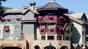 Um edifício de vários andares com fachada de pedra, várias varandas com floreiras e uma entrada destacada com arco de pedra.