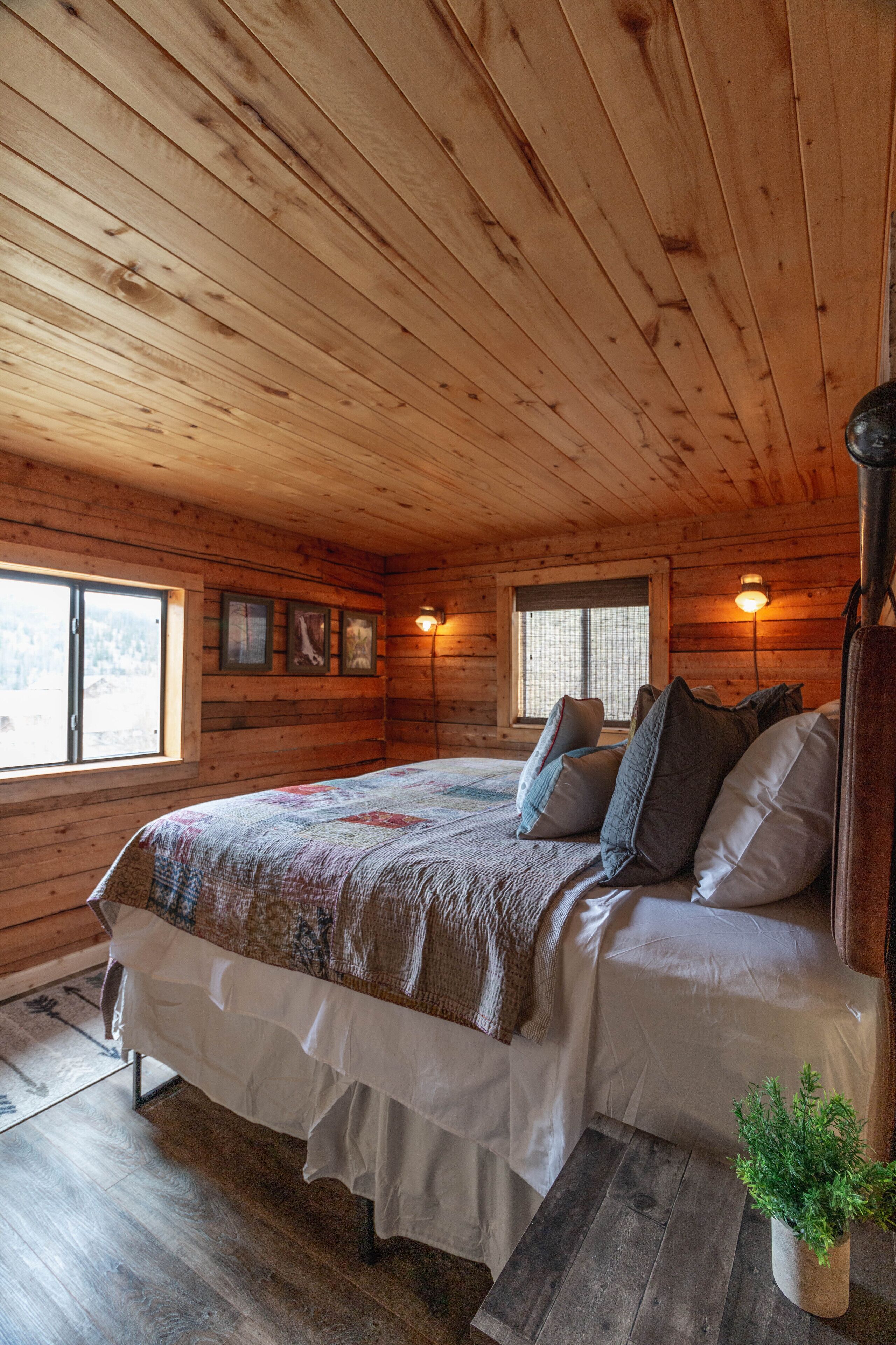 Ein Schlafzimmer einer Holzhütte mit einem Bett, zwei Fenstern, einer Wandleuchte und einem kleinen Topfpflanzen.