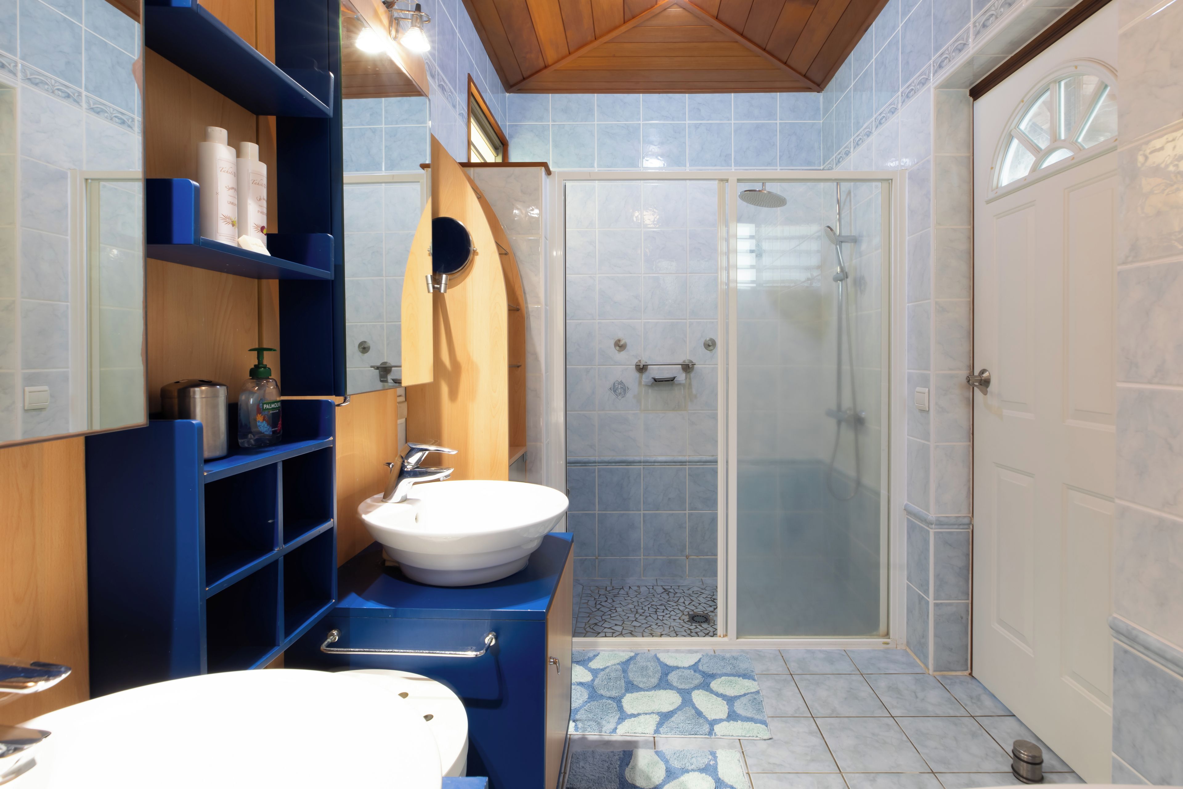 Un baño con lavamanos blanco, estantes de madera, un espejo y una ducha con mampara de vidrio.