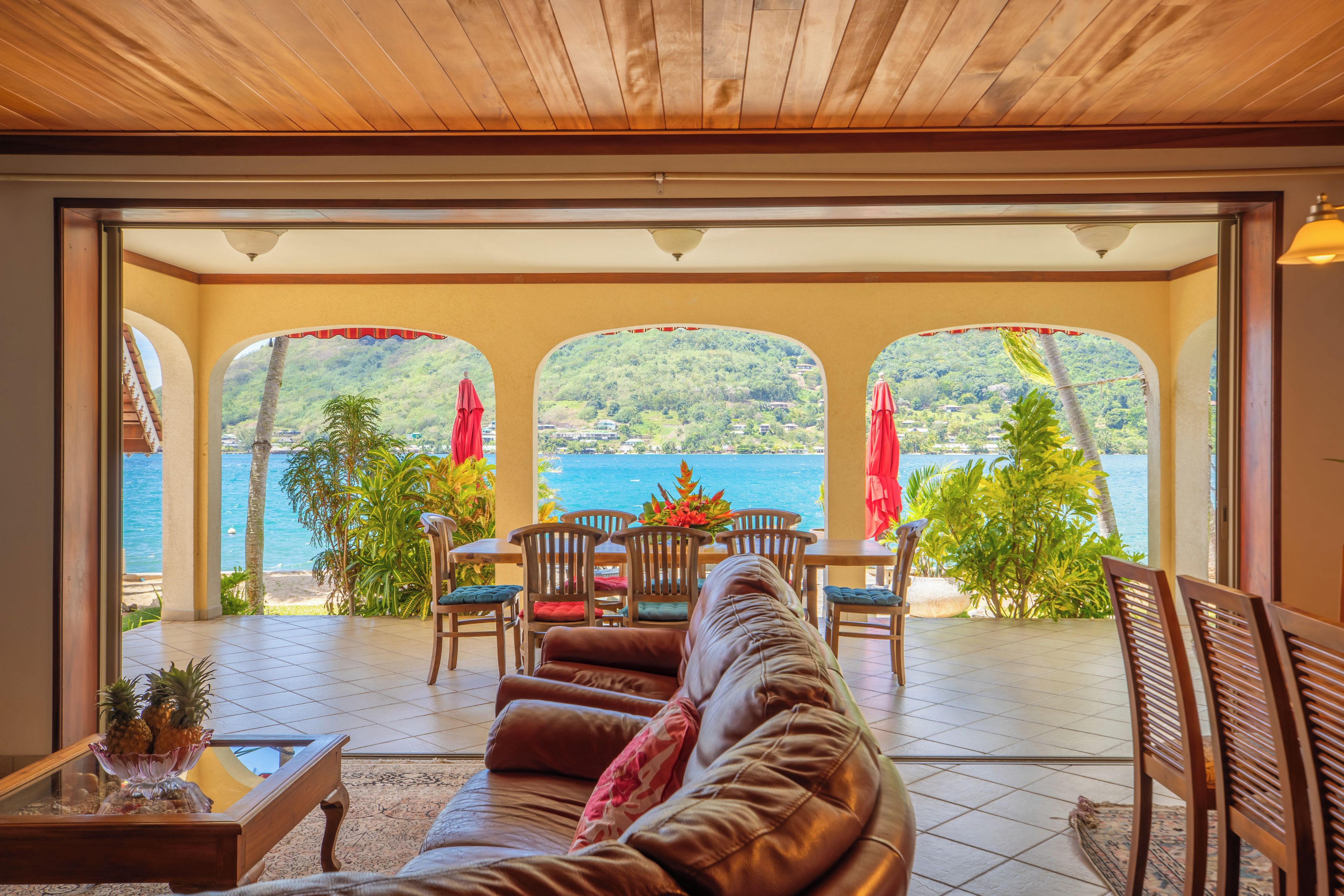 A spacious patio with a leather sofa, wooden chairs, and a dining table. It features a view of a body of water and lush greenery.