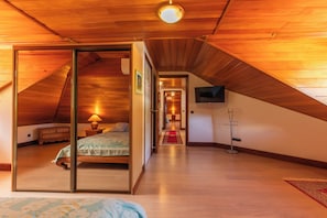 A room with a sloped ceiling, a bed with a nightstand, a lamp, and a television mounted on the wall.