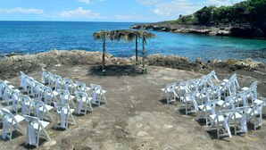 An outdoor wedding setup with white chairs arranged in rows, a small altar with a palm tree, and a rocky shoreline with clear blue water. - Magical Romantic Getaway For 2, Lovers Paradise On Private White Sand Beach Cove (St Mary)