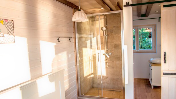 Dusche mit Glastüren, Holzbalken an der Decke, ein Fenster mit Blick auf Grünflächen und ein weißes Waschbecken.