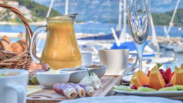 A table set with breakfast items including bread, cheese, and fruit, with a view of boats and mountains in the background.