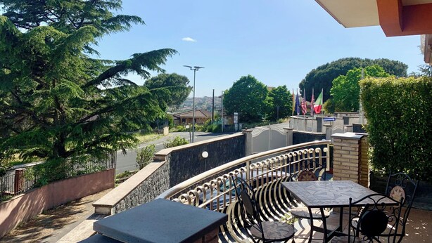 Eine Terrasse mit Sitzgelegenheit im Freien, Blick auf eine Straße und ein Gebäude mit Flaggen im Hintergrund.