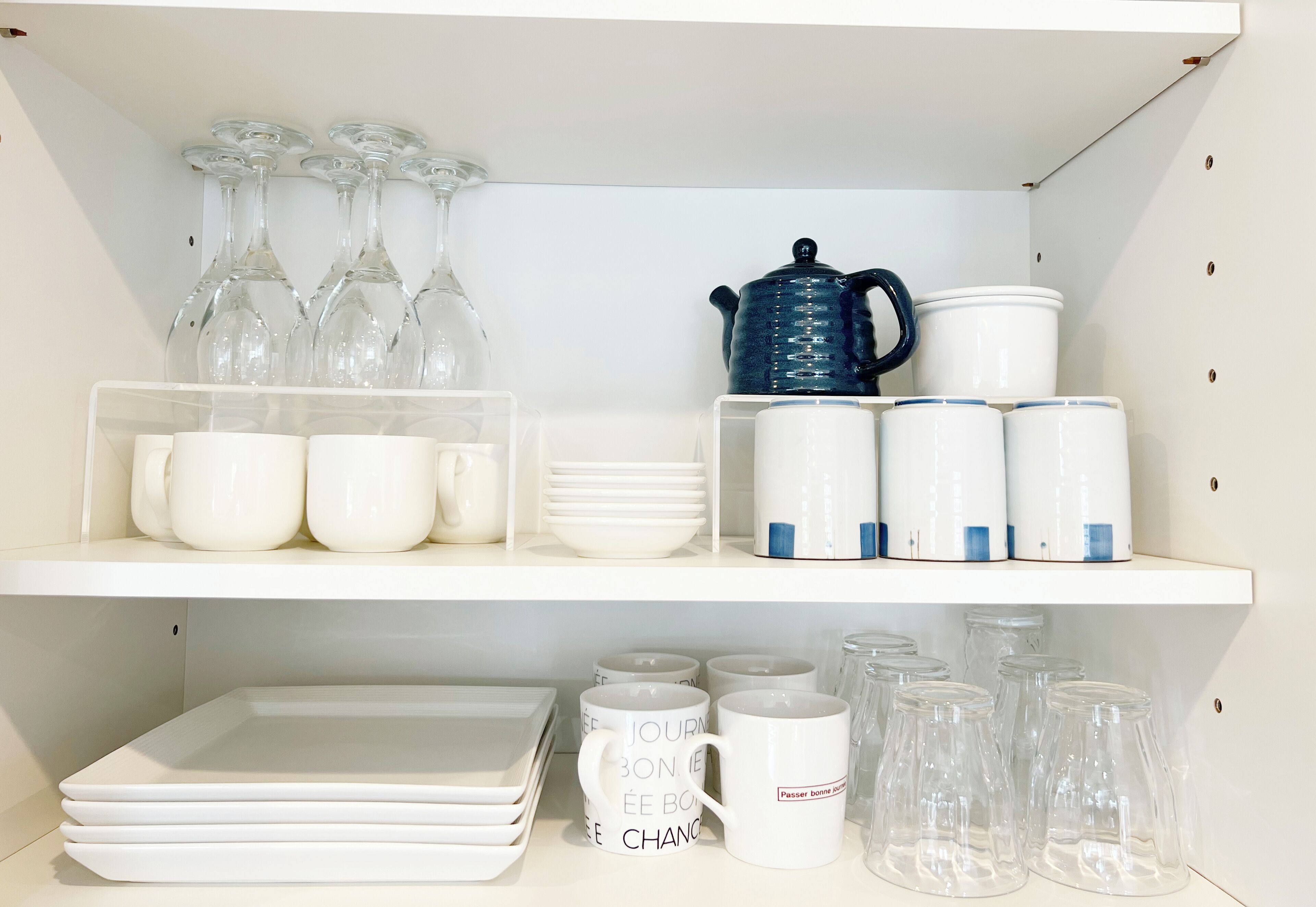 A kitchen shelf with white and blue ceramic mugs, clear glass cups, and a blue teapot.