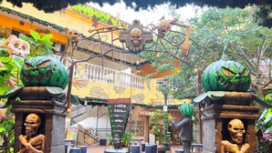 Een buitenplaats met decoratieve skeletten, een groene pompoen en een metalen boog met borden voor verschillende winkels.