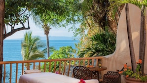 A balcony with a bed, a table, and chairs, overlooking a body of water and surrounded by greenery. - Villa Estrella Mar - 5 Star Luxury Villa - Staff - Private Pool - On the beach! (Puerto Vallarta)