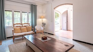 A room with a wooden bench, a white chair, a table with a decorative box, a floor lamp, and a view of the outside through a window with curtains. - Costabravaforrent Tallada (La Tallada d'Empordà)